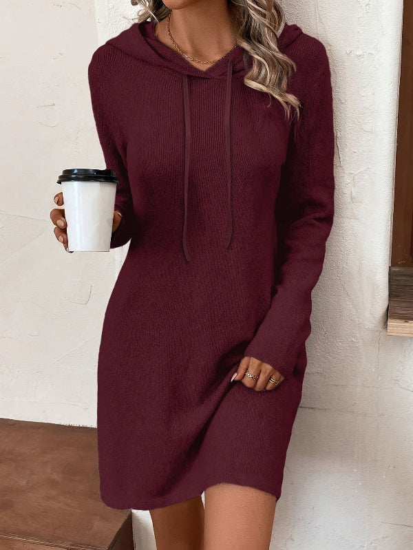 Person wearing a burgundy hoodie dress holding a coffee cup against a white brick wall.