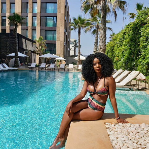 Woman in a colorful bikini sitting by a pool with palm trees and a building in the background